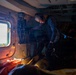 The image shows the interior of a military or search-and-rescue helicopter with at least two personnel visible. One individual wearing a camouflage uniform and communications headset is leaning over what appears to be equipment or a casualty on the floor. The scene is dimly lit with warm orange lighting accents, suggesting an active operational mission.