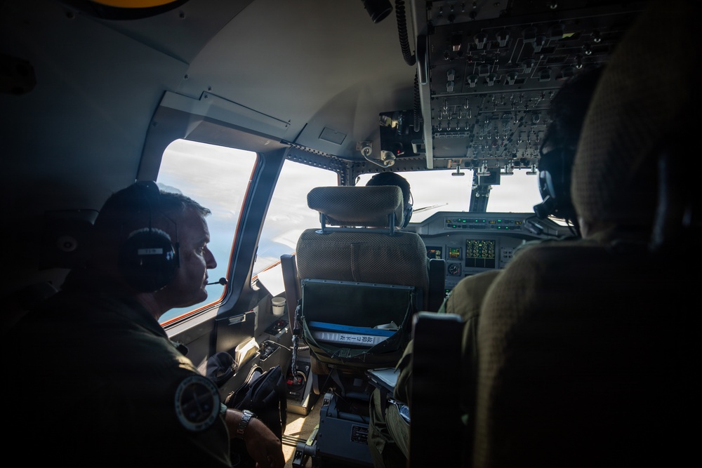 The image shows the interior of a military aircraft cockpit with at least two crew members visible, both wearing aviation headsets and olive/flight suits. The aircraft appears to be in flight, with clouds visible through the cockpit windows. The cockpit features multiple instrument panels, overhead control panels, and modern avionics displays.