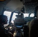 The image shows the interior of a military aircraft cockpit with at least two crew members visible, both wearing aviation headsets and olive/flight suits. The aircraft appears to be in flight, with clouds visible through the cockpit windows. The cockpit features multiple instrument panels, overhead control panels, and modern avionics displays.