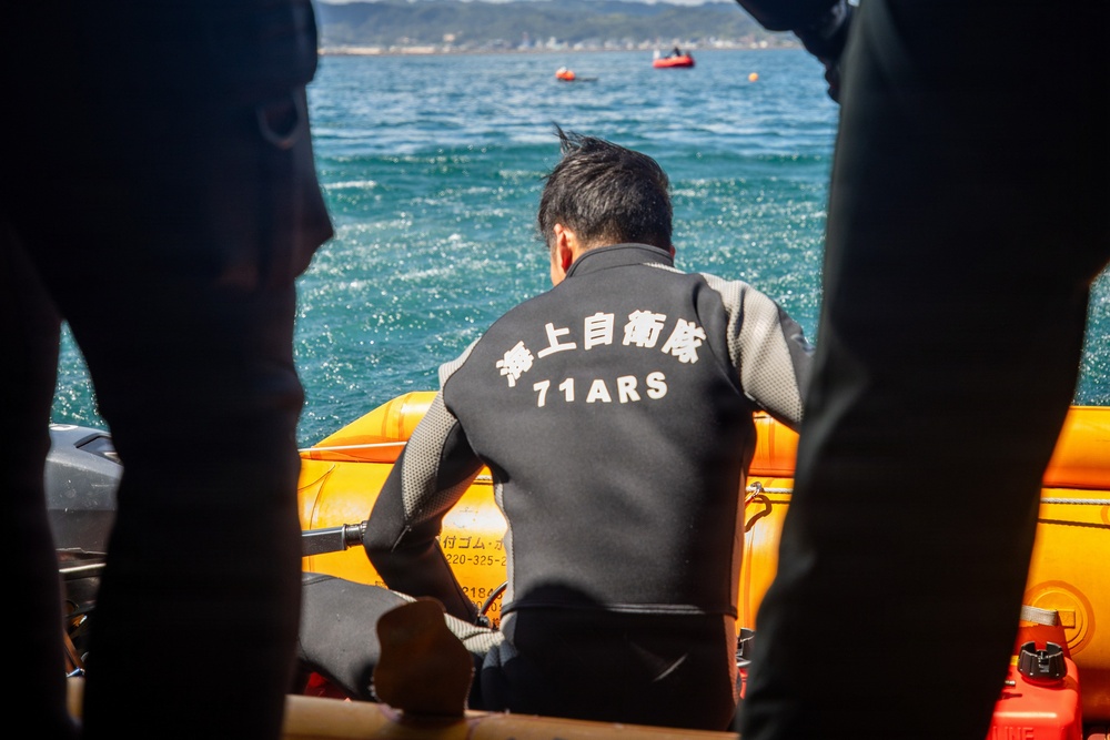 A person wearing a black wetsuit marked with Japanese characters '海上自衛隊' (Japan Maritime Self-Defense Force) and '71ARS' is seated on a yellow inflatable rescue boat, facing the open sea. The scene appears to be a maritime rescue or training operation. Other individuals in wetsuits are partially visible in the foreground, framing the central figure.