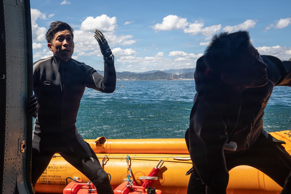 Two individuals wearing black wetsuits are aboard an orange inflatable rescue/dive boat on open water. One person faces the camera with a raised gloved hand, appearing to communicate or signal, while the other is bent over or adjusting equipment. A coastline with mountains and what appears to be an industrial facility is visible in the background.