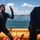 U.S. Marines, JMSDF Sailors reinforce maritime readiness during Rescue Flag 26-1 Two individuals wearing black wetsuits are aboard an orange inflatable rescue/dive boat on open water. One person faces the camera with a raised gloved hand, appearing to communicate or signal, while the other is bent over or adjusting equipment. A coastline with mountains and what appears to be an industrial facility is visible in the background.