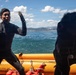 Two individuals wearing black wetsuits are aboard an orange inflatable rescue/dive boat on open water. One person faces the camera with a raised gloved hand, appearing to communicate or signal, while the other is bent over or adjusting equipment. A coastline with mountains and what appears to be an industrial facility is visible in the background.