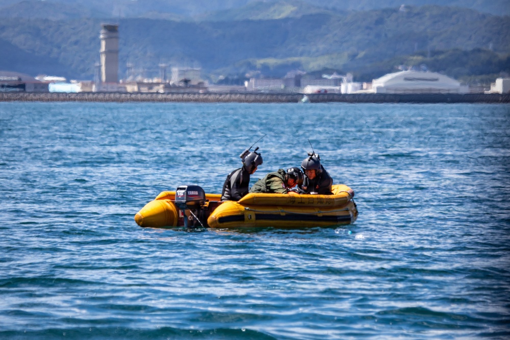A yellow inflatable rubber dinghy with a Yamaha 15 outboard motor is operating on open blue water. Three individuals wearing helmets and dark gear are aboard, with one appearing to be lying down or being assisted, suggesting a rescue or training operation. An industrial waterfront with a tall tower structure and green hills is visible in the background.