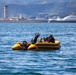 A yellow inflatable rubber dinghy with a Yamaha 15 outboard motor is operating on open blue water. Three individuals wearing helmets and dark gear are aboard, with one appearing to be lying down or being assisted, suggesting a rescue or training operation. An industrial waterfront with a tall tower structure and green hills is visible in the background.