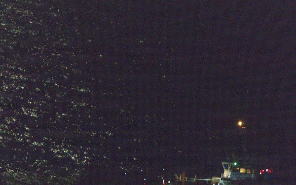 A nighttime maritime scene showing a vessel illuminated by onboard lights near what appears to be a rocky or vegetated shoreline. The water surface reflects ambient light on the left side of the frame. Overall visibility is very limited due to darkness.