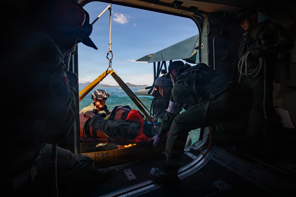 A helicopter rescue operation is underway over open water, with crew members hoisting an injured or incapacitated person on a rescue litter/stretcher into the aircraft. Multiple personnel in flight suits and helmets are coordinating the extraction, with a hoist cable and rigging system visible. The scene appears to be a maritime search and rescue exercise or actual rescue operation, with mountainous coastline visible in the background.