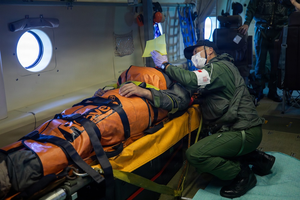 A medical personnel in military/rescue uniform is attending to a patient secured in an orange rescue/evacuation bag on a stretcher inside what appears to be a military or coast guard aircraft. The attendant is wearing a face mask, gloves, and a green flight suit with a red cross armband, and is reviewing what appears to be a yellow medical document. The setting suggests an aeromedical evacuation or search-and-rescue operation.