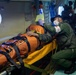 A medical personnel in military/rescue uniform is attending to a patient secured in an orange rescue/evacuation bag on a stretcher inside what appears to be a military or coast guard aircraft. The attendant is wearing a face mask, gloves, and a green flight suit with a red cross armband, and is reviewing what appears to be a yellow medical document. The setting suggests an aeromedical evacuation or search-and-rescue operation.