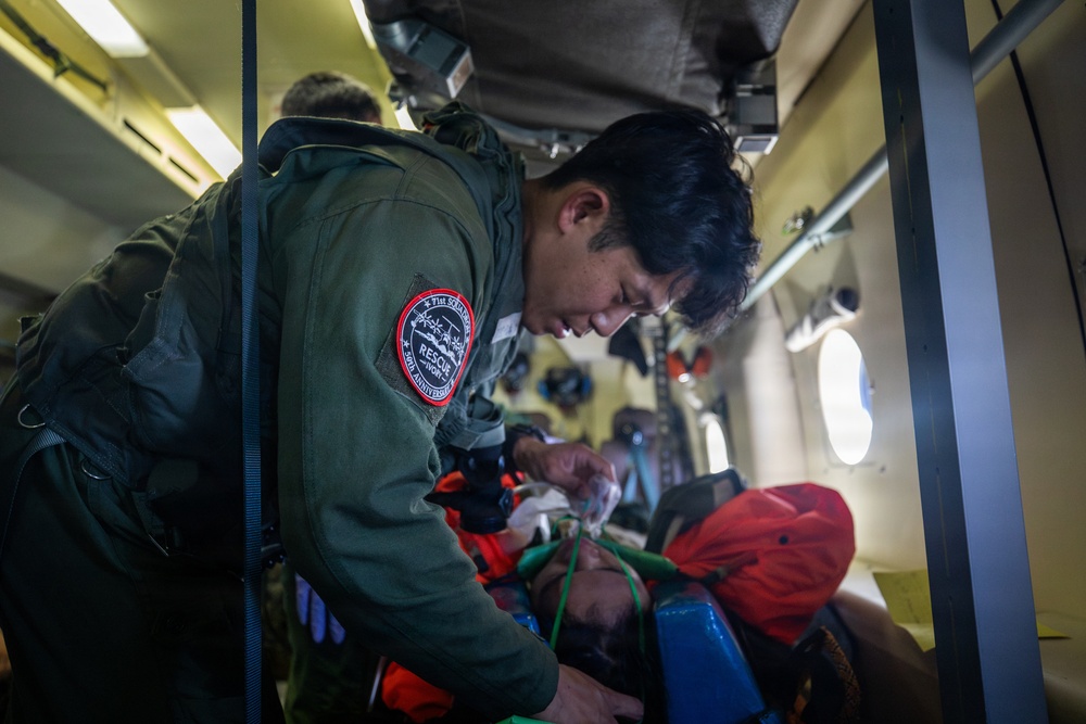 A rescue personnel wearing a military-style flight suit with a 'Rescue' unit patch is attending to a patient lying on a stretcher inside what appears to be a military or rescue helicopter. The patient is wearing an orange jacket and has an oxygen mask applied to their face. The scene depicts an active aeromedical evacuation or search-and-rescue operation in progress.