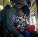 A rescue personnel wearing a military-style flight suit with a 'Rescue' unit patch is attending to a patient lying on a stretcher inside what appears to be a military or rescue helicopter. The patient is wearing an orange jacket and has an oxygen mask applied to their face. The scene depicts an active aeromedical evacuation or search-and-rescue operation in progress.