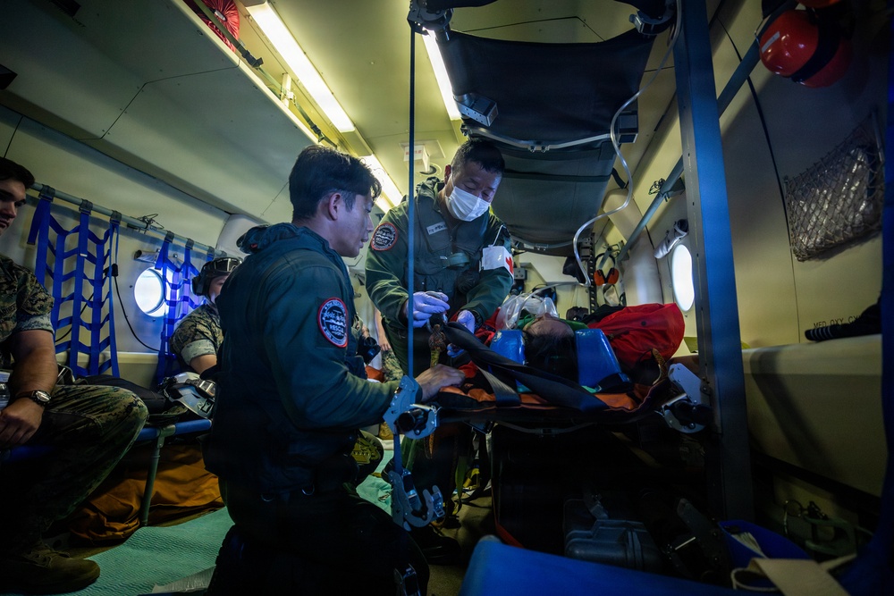 The image shows a medical evacuation scenario inside what appears to be a military aircraft or helicopter. Two personnel in flight suits are attending to a patient on a stretcher, with additional military personnel visible in the background. The setting suggests an aeromedical evacuation or MEDEVAC training or operational mission.