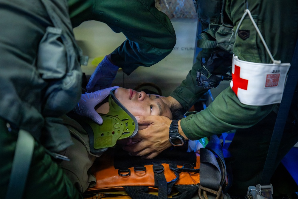 Military medical personnel are providing emergency care to a casualty secured on an orange stretcher. Two individuals in olive/green flight suits are attending to a patient who has a cervical collar applied, with one wearing blue medical gloves. A red cross medical pouch is visible on one responder's arm, indicating this is a military medical or combat casualty care scenario.
