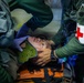 Military medical personnel are providing emergency care to a casualty secured on an orange stretcher. Two individuals in olive/green flight suits are attending to a patient who has a cervical collar applied, with one wearing blue medical gloves. A red cross medical pouch is visible on one responder's arm, indicating this is a military medical or combat casualty care scenario.