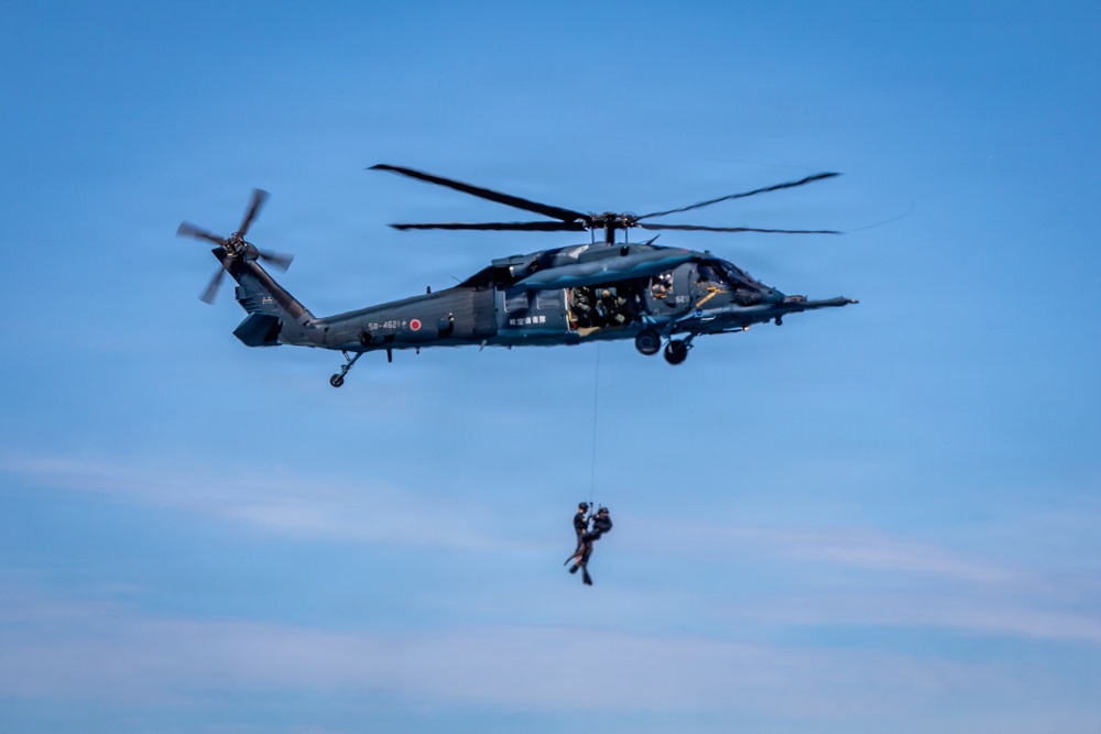 A dark gray military helicopter, identifiable as a Japan Air Self-Defense Force UH-60 Black Hawk variant, hovers against a clear blue sky. A person in tactical gear is suspended below the aircraft on a hoist or rappel line. The scene appears to depict a combat search and rescue (CSAR) or special operations training exercise.