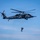 U.S. Marines, JMSDF Sailors reinforce maritime readiness during Rescue Flag 26-1 A dark gray military helicopter, identifiable as a Japan Air Self-Defense Force UH-60 Black Hawk variant, hovers against a clear blue sky. A person in tactical gear is suspended below the aircraft on a hoist or rappel line. The scene appears to depict a combat search and rescue (CSAR) or special operations training exercise.