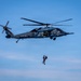 A dark gray military helicopter, identifiable as a Japan Air Self-Defense Force UH-60 Black Hawk variant, hovers against a clear blue sky. A person in tactical gear is suspended below the aircraft on a hoist or rappel line. The scene appears to depict a combat search and rescue (CSAR) or special operations training exercise.