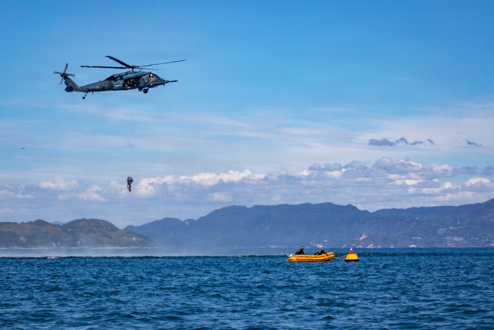 A military or coast guard helicopter is conducting a water rescue or training operation over open sea. A person is being hoisted or lowered on a cable beneath the helicopter, while two yellow inflatable rafts with personnel are positioned on the water below. Mountains and coastline are visible in the background under partly cloudy skies.