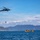 U.S. Marines, JMSDF Sailors reinforce maritime readiness during Rescue Flag 26-1 A military or coast guard helicopter is conducting a water rescue or training operation over open sea. A person is being hoisted or lowered on a cable beneath the helicopter, while two yellow inflatable rafts with personnel are positioned on the water below. Mountains and coastline are visible in the background under partly cloudy skies.