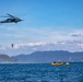 A military or coast guard helicopter is conducting a water rescue or training operation over open sea. A person is being hoisted or lowered on a cable beneath the helicopter, while two yellow inflatable rafts with personnel are positioned on the water below. Mountains and coastline are visible in the background under partly cloudy skies.