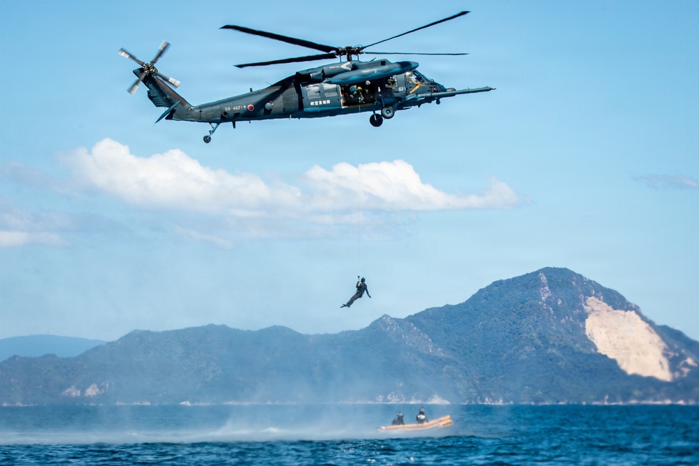 A military helicopter, identifiable as a Sikorsky SH-60 (Black Hawk variant) with Japanese markings, hovers over open water while a person rappels or is being lowered/dropped toward a rigid inflatable boat (RIB) below. The scene appears to be a military training exercise or rescue operation conducted in coastal waters with mountainous islands visible in the background. The operation involves coordination between the airborne crew and personnel on the watercraft.