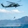 U.S. Marines, JMSDF Sailors reinforce maritime readiness during Rescue Flag 26-1 A military helicopter, identifiable as a Sikorsky SH-60 (Black Hawk variant) with Japanese markings, hovers over open water while a person rappels or is being lowered/dropped toward a rigid inflatable boat (RIB) below. The scene appears to be a military training exercise or rescue operation conducted in coastal waters with mountainous islands visible in the background. The operation involves coordination between the airborne crew and personnel on the watercraft.