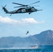 A military helicopter, identifiable as a Sikorsky SH-60 (Black Hawk variant) with Japanese markings, hovers over open water while a person rappels or is being lowered/dropped toward a rigid inflatable boat (RIB) below. The scene appears to be a military training exercise or rescue operation conducted in coastal waters with mountainous islands visible in the background. The operation involves coordination between the airborne crew and personnel on the watercraft.