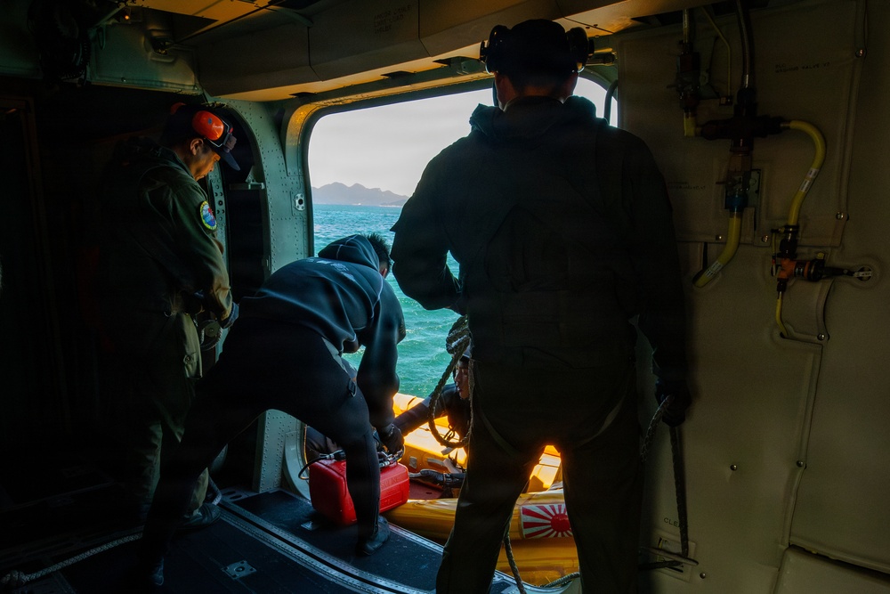The image shows the interior of a military helicopter with personnel conducting what appears to be a maritime rescue or search-and-rescue operation. Three individuals are visible near the open side door of the aircraft, with one person in a wetsuit handling equipment on a yellow rescue raft or platform. A Japanese Maritime Self-Defense Force rising sun emblem is visible on equipment in the foreground.