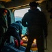 The image shows the interior of a military helicopter with personnel conducting what appears to be a maritime rescue or search-and-rescue operation. Three individuals are visible near the open side door of the aircraft, with one person in a wetsuit handling equipment on a yellow rescue raft or platform. A Japanese Maritime Self-Defense Force rising sun emblem is visible on equipment in the foreground.