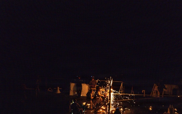 A nighttime scene on what appears to be a ship's deck or industrial platform, where workers are engaged in welding or cutting operations producing bright orange sparks. The image is very dark overall with the work area illuminated primarily by the sparks and small ambient lights. Multiple personnel are visible around the work site.