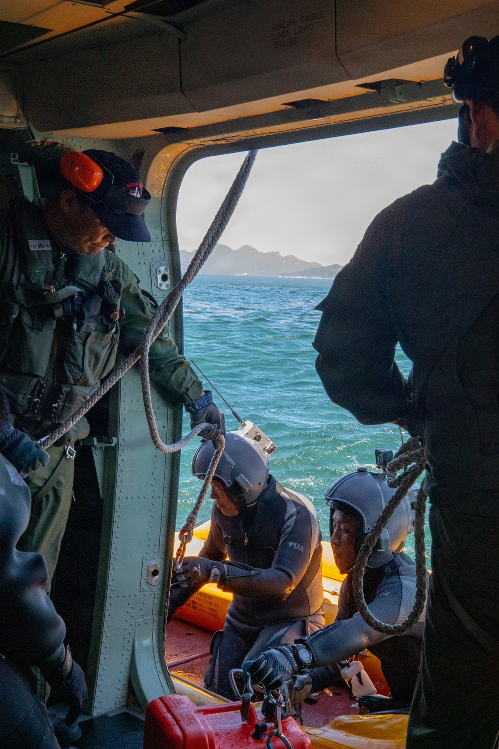 The image shows what appears to be a maritime rescue or military operation, with personnel inside a helicopter or vessel doorway interacting with two divers in wetsuits and helmets on a yellow inflatable rescue boat. A crew member in olive/green flight suit with orange ear protection is managing ropes, while two wetsuit-clad divers with helmets are positioned on the bright yellow rigid inflatable boat (RIB). The scene takes place over open ocean water with mountainous coastline visible in the background.