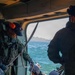 The image shows what appears to be a maritime rescue or military operation, with personnel inside a helicopter or vessel doorway interacting with two divers in wetsuits and helmets on a yellow inflatable rescue boat. A crew member in olive/green flight suit with orange ear protection is managing ropes, while two wetsuit-clad divers with helmets are positioned on the bright yellow rigid inflatable boat (RIB). The scene takes place over open ocean water with mountainous coastline visible in the background.