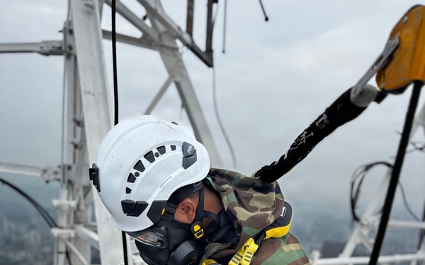 A worker in full personal protective equipment is positioned at height on a white metal tower or antenna structure. They are wearing camouflage clothing, a white climbing helmet, a full-face respirator mask, and an elaborate yellow-and-black fall arrest harness system with multiple carabiners. The background reveals an elevated urban or semi-urban landscape under overcast skies, indicating significant working height.