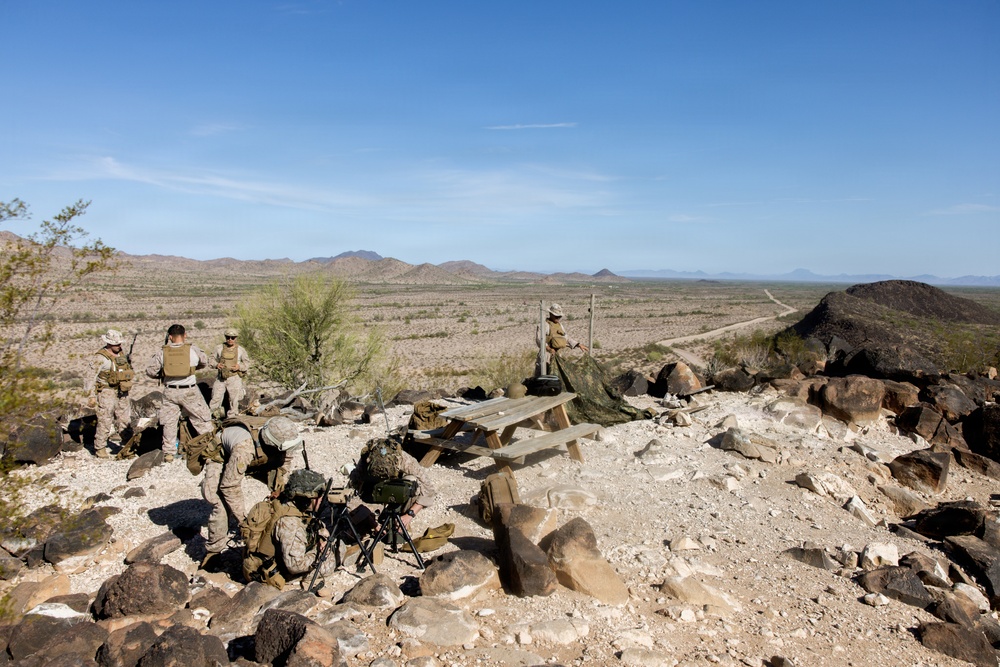 A group of military personnel in desert camouflage uniforms are positioned on a rocky hilltop overlooking a vast arid desert landscape with distant mountains. The soldiers appear to be conducting a training exercise or observation post operation, with equipment including what appears to be a tripod-mounted device. A wooden picnic table and camouflage netting are also visible at the elevated position.