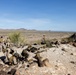 A group of military personnel in desert camouflage uniforms are positioned on a rocky hilltop overlooking a vast arid desert landscape with distant mountains. The soldiers appear to be conducting a training exercise or observation post operation, with equipment including what appears to be a tripod-mounted device. A wooden picnic table and camouflage netting are also visible at the elevated position.