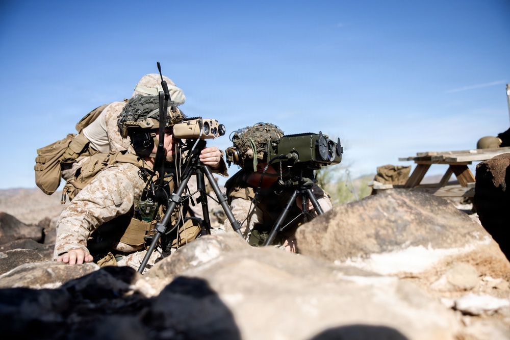 Two military personnel in desert camouflage uniforms are positioned behind rocks in an arid environment, operating optical/surveillance equipment mounted on tripods. The setting appears to be a desert training range, with clear blue skies and rocky terrain visible. Both individuals are in a low-profile observation posture, suggesting a forward observer or reconnaissance exercise.