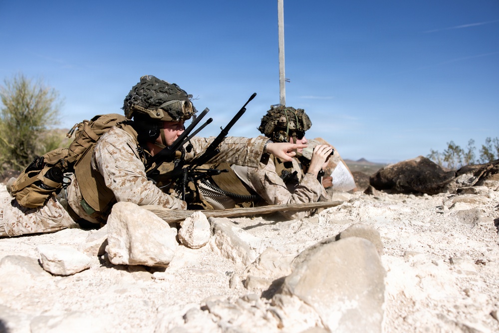 Two military personnel in desert camouflage uniforms are in a prone position among rocky terrain, appearing to be in a tactical training exercise. The soldier on the left is operating what appears to be a machine gun with an ammunition belt, while pointing toward a target or objective. The setting is a arid, rocky desert landscape under clear blue skies.