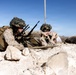 Two military personnel in desert camouflage uniforms are in a prone position among rocky terrain, appearing to be in a tactical training exercise. The soldier on the left is operating what appears to be a machine gun with an ammunition belt, while pointing toward a target or objective. The setting is a arid, rocky desert landscape under clear blue skies.