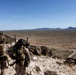 Two military personnel in full combat gear are positioned on a rocky elevated outcrop overlooking a vast desert landscape. One soldier appears to be pointing toward the terrain below, suggesting reconnaissance or tactical observation activity. The setting appears to be an arid desert environment, consistent with a training exercise or operational deployment.