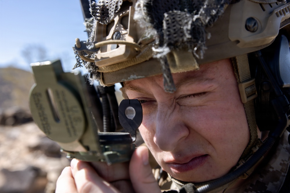 A close-up photograph of a military service member using a lensatic compass to take a bearing or azimuth reading. The individual is wearing a tan/coyote brown tactical helmet with netting, communications equipment, and digital camouflage uniform. The background shows a rocky, arid terrain under a clear blue sky.