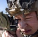 A close-up photograph of a military service member using a lensatic compass to take a bearing or azimuth reading. The individual is wearing a tan/coyote brown tactical helmet with netting, communications equipment, and digital camouflage uniform. The background shows a rocky, arid terrain under a clear blue sky.