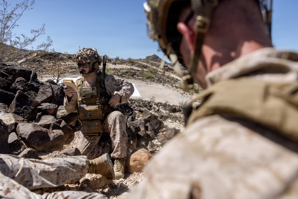 Two military personnel in desert camouflage uniforms are positioned among rocks in an arid, rocky terrain under a clear blue sky. The soldier in focus is seated among dark rocks, reviewing what appears to be a notepad or small document while wearing full tactical gear including a helmet with communications equipment. A second soldier is visible in the foreground, out of focus, also in full combat kit.