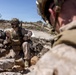 Two military personnel in desert camouflage uniforms are positioned among rocks in an arid, rocky terrain under a clear blue sky. The soldier in focus is seated among dark rocks, reviewing what appears to be a notepad or small document while wearing full tactical gear including a helmet with communications equipment. A second soldier is visible in the foreground, out of focus, also in full combat kit.