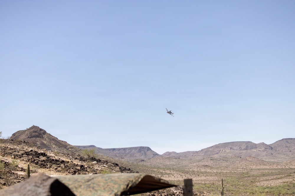 A military or fixed-wing aircraft is captured in flight over a desert landscape, likely in the American Southwest based on the terrain and vegetation. The scene shows arid, rocky mountains in the background with sparse desert scrub and saguaro cacti visible. A camouflage-patterned object, possibly military equipment or netting, is partially visible in the foreground.