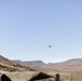 A military or fixed-wing aircraft is captured in flight over a desert landscape, likely in the American Southwest based on the terrain and vegetation. The scene shows arid, rocky mountains in the background with sparse desert scrub and saguaro cacti visible. A camouflage-patterned object, possibly military equipment or netting, is partially visible in the foreground.
