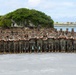 A large group of military personnel, approximately 50-60 individuals, pose for a formal unit photograph in two rows with arms crossed. They are wearing matching woodland camouflage uniforms (MARPAT pattern) consistent with U.S. Marine Corps dress, standing on a paved surface near a coastal waterfront. A distinctive rocky outcropping with trees growing on top serves as the backdrop, suggesting a Pacific island location.