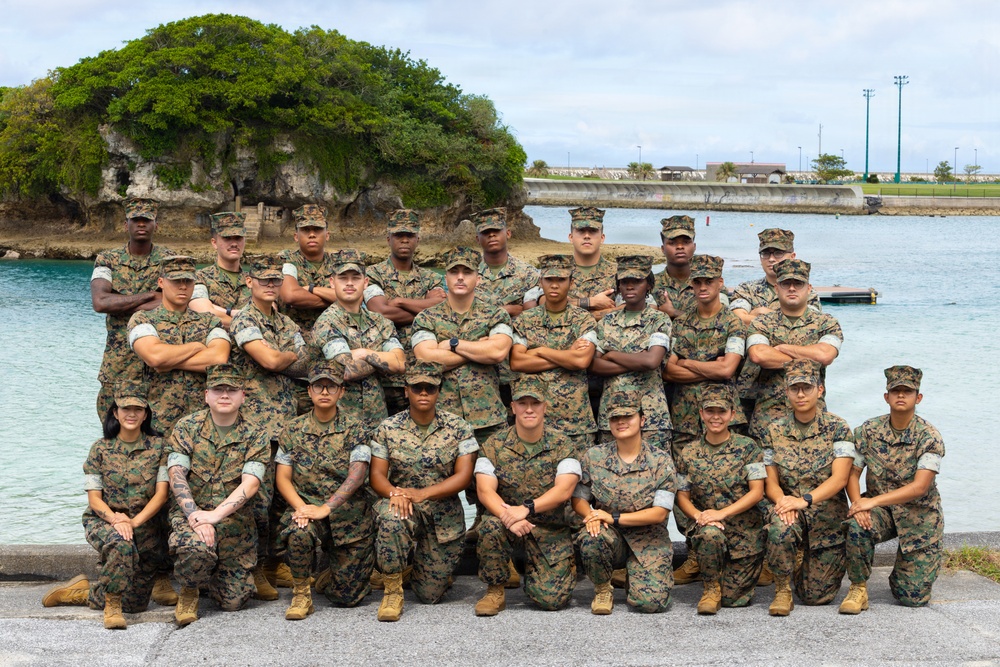 A group of approximately 25-30 military personnel in U.S. Marine Corps MARPAT (woodland digital camouflage) uniforms pose for a formal group photograph. They are arranged in three rows — back row standing, middle row with arms crossed, and front row kneeling — in front of a coastal waterway with a rocky limestone outcropping and tree in the background. The setting appears to be a military installation near a body of water, consistent with locations in the Pacific region such as Okinawa or Guam.