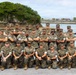 A group of approximately 25-30 military personnel in U.S. Marine Corps MARPAT (woodland digital camouflage) uniforms pose for a formal group photograph. They are arranged in three rows — back row standing, middle row with arms crossed, and front row kneeling — in front of a coastal waterway with a rocky limestone outcropping and tree in the background. The setting appears to be a military installation near a body of water, consistent with locations in the Pacific region such as Okinawa or Guam.