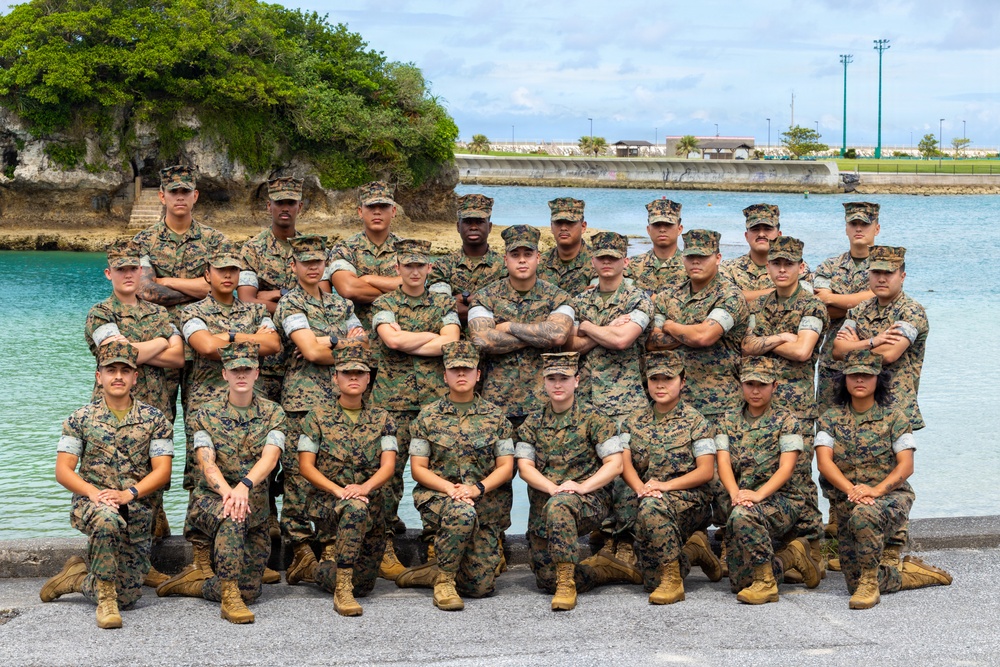 A group of approximately 25-30 U.S. Marines in woodland MARPAT camouflage uniforms pose for a formal unit photograph near a waterfront location. The personnel are arranged in three rows — front row kneeling, middle row standing with arms crossed, and back row standing — in a classic military group portrait formation. The setting appears to be a coastal military installation, likely in a Pacific region based on the tropical vegetation and turquoise water visible in the background.