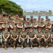 A group of approximately 25-30 U.S. Marines in woodland MARPAT camouflage uniforms pose for a formal unit photograph near a waterfront location. The personnel are arranged in three rows — front row kneeling, middle row standing with arms crossed, and back row standing — in a classic military group portrait formation. The setting appears to be a coastal military installation, likely in a Pacific region based on the tropical vegetation and turquoise water visible in the background.